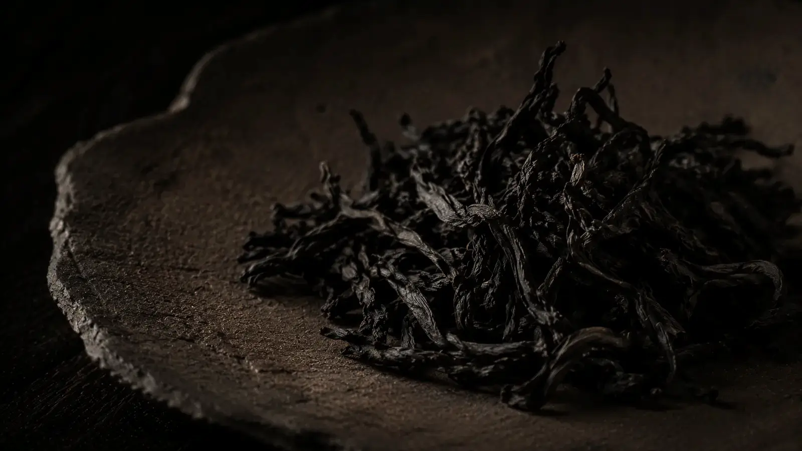 Dark roasted Da Hong Pao oolong leaves on bamboo scoop beside clay teapot on stone, evoking Wuyi Mountain terroir.