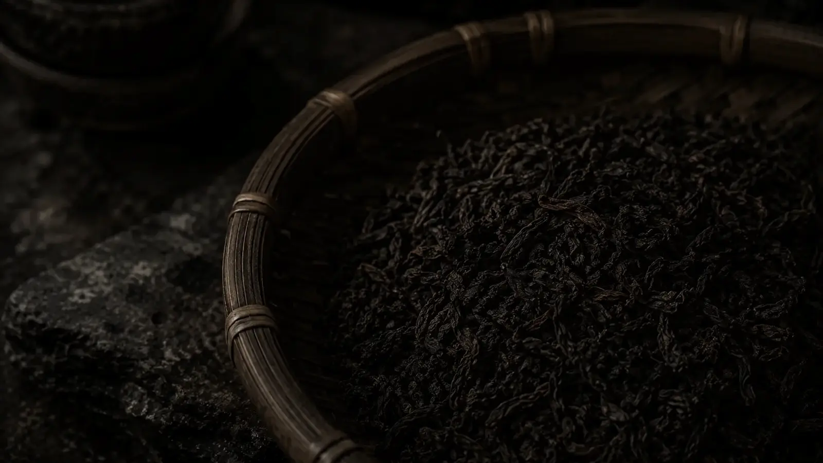 Dark aged Liu Bao tea leaves spilling from a bamboo basket onto stone, beside a cup of deep reddish-brown brewed tea.