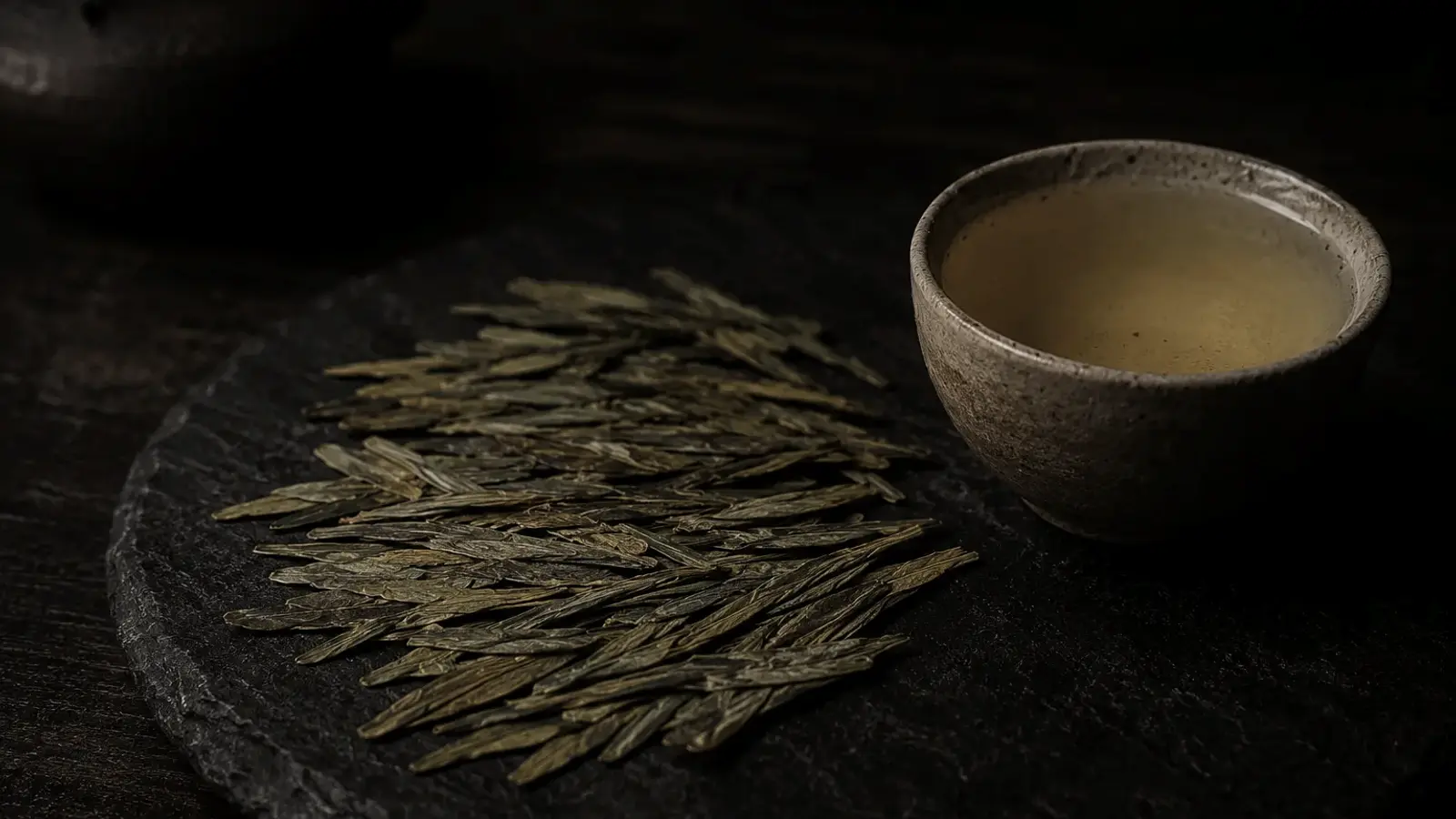 Flat-pressed Longjing tea leaves beside a clear glass of pale jade-green liquor with upright leaves, brewed in natural light.