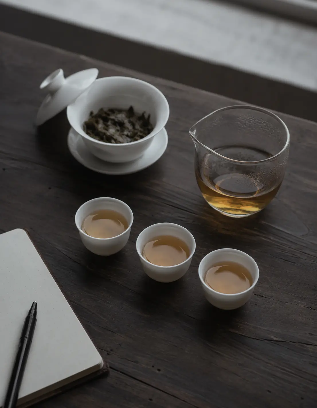 Gongfu tea tasting session with gaiwan, fairness pitcher, cups, and tasting notebook on dark wood